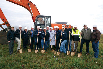 A group of people standing in the grass holding shovels in front of a construction machine